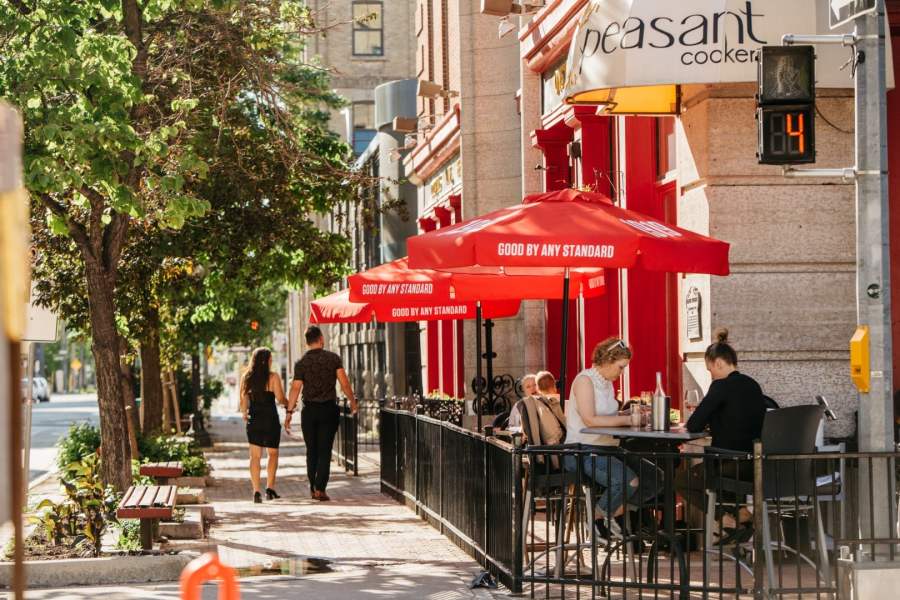 A sidewalk scene in the Exchange District featuring a café with red umbrellas labeled 'Good by Any Standard'. Two people walk hand in hand while two others dine at a patio.