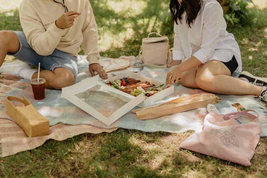Two people enjoying a picnic on a blanket in a grassy area, surrounded by food items in boxes and bags.