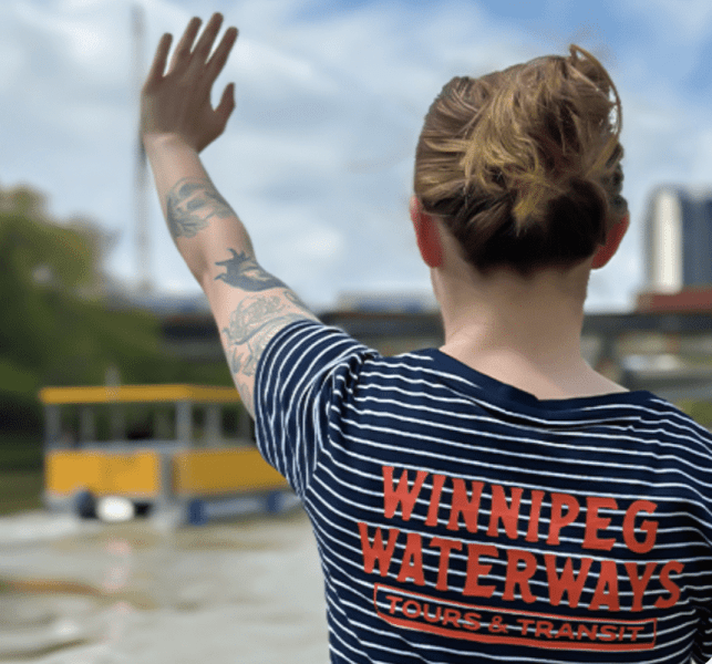 A person wearing a striped shirt with 'Winnipeg Waterways Tours & Transit' on the back, standing by the water and waving towards a small yellow boat in the background.