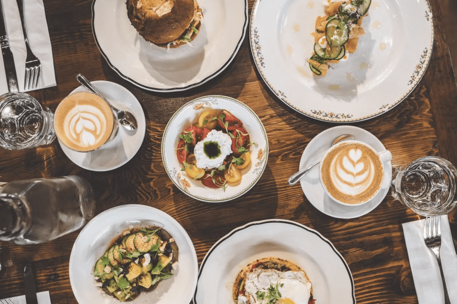 A top-down view of a brunch spread featuring a variety of dishes, including a sandwich, a colorful salad with cherry tomatoes, a bowl of roasted vegetables, and two cups of coffee with latte art on a wooden table.