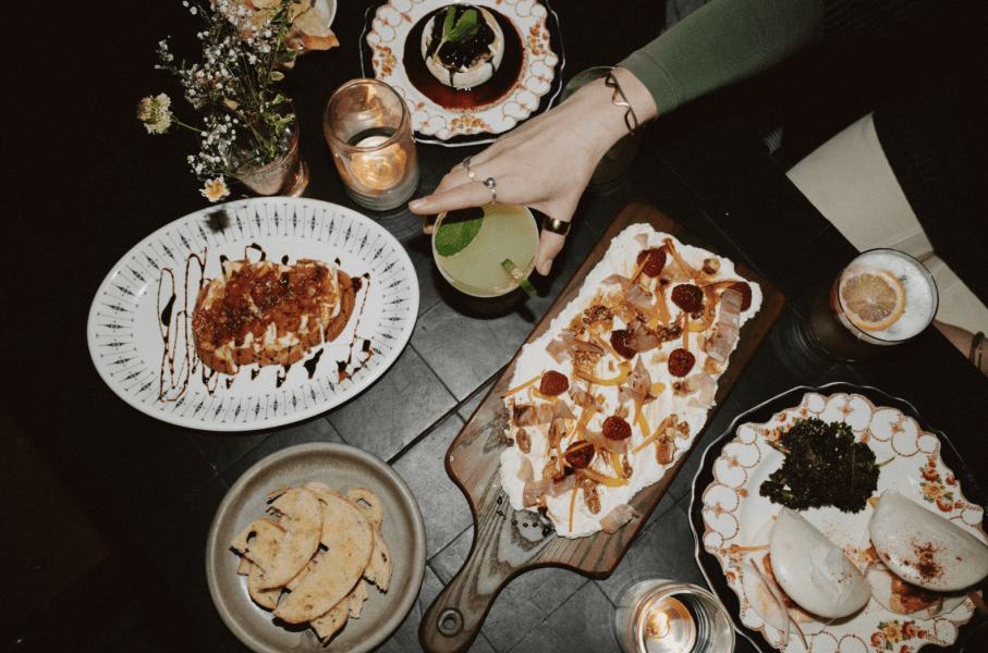 A flat lay of a dining table featuring various dishes, including a plate of bruschetta with toppings, a dessert, a platter with creamy cheese and fruits, and cocktails, surrounded by candles and flowers.