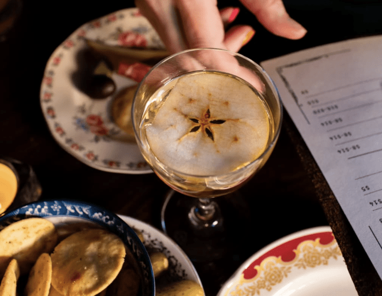 A close-up image of a cocktail garnished with a star-patterned slice of apple, surrounded by various appetizers on decorative plates, with a menu partially visible in the background.