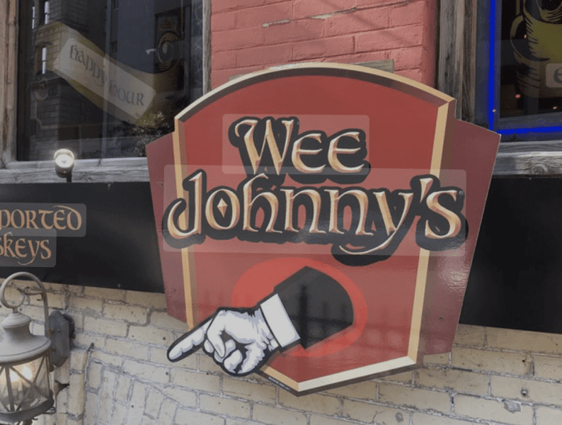 Sign for Wee Johnny's Irish Pub featuring a hand pointing, set against a red and black background on a brick wall.