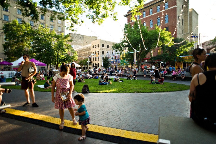 A vibrant outdoor scene at the Winnipeg International Fringe Festival, featuring families and individuals enjoying the festivities on the grass in the Exchange District, with food vendors and colorful decorations in the background.