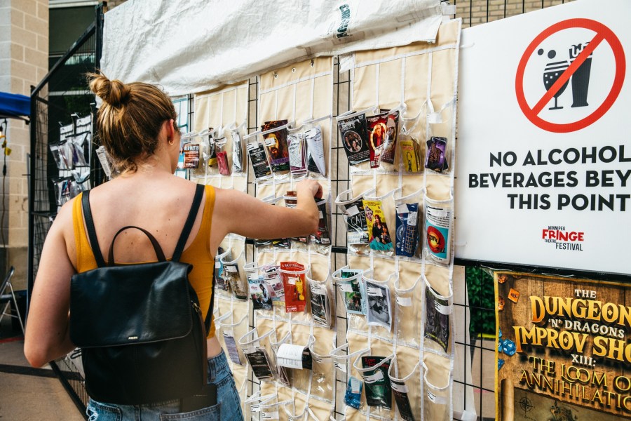 A person with a backpack examines a display of festival pamphlets at the Winnipeg International Fringe Festival, with a sign indicating no alcohol beverages beyond that point.