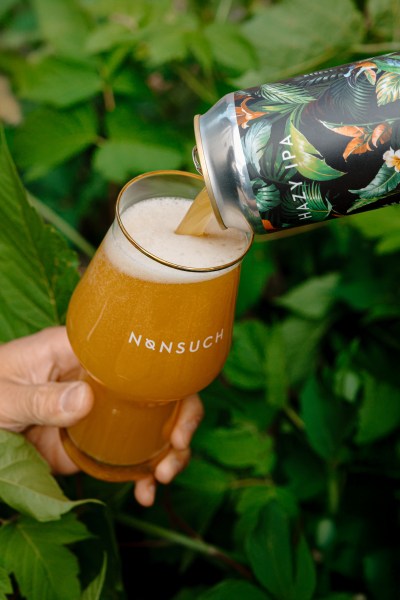 A hand holding a glass with the Nonsuch logo, filled with a hazy IPA beer, while pouring from a can decorated with tropical foliage. Background features green leaves.