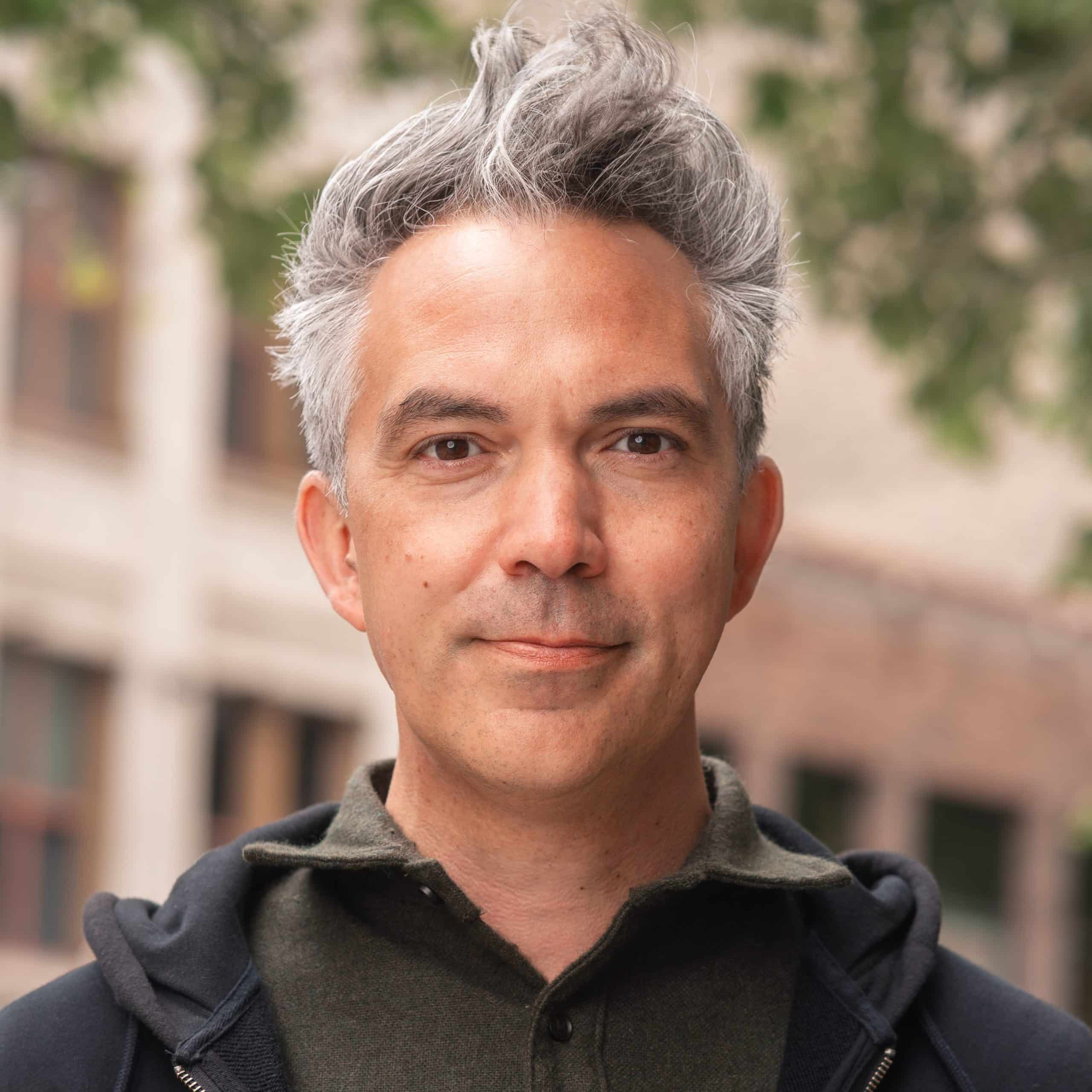 Portrait of a man with gray hair wearing a dark hoodie and button-up shirt, standing outdoors with a blurred building in the background.