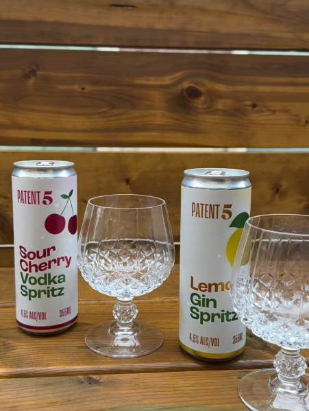 Two cans of Patent 5 spritz drinks, one Sour Cherry Vodka Spritz and one Lemon Gin Spritz, placed next to crystal glasses on a wooden surface.