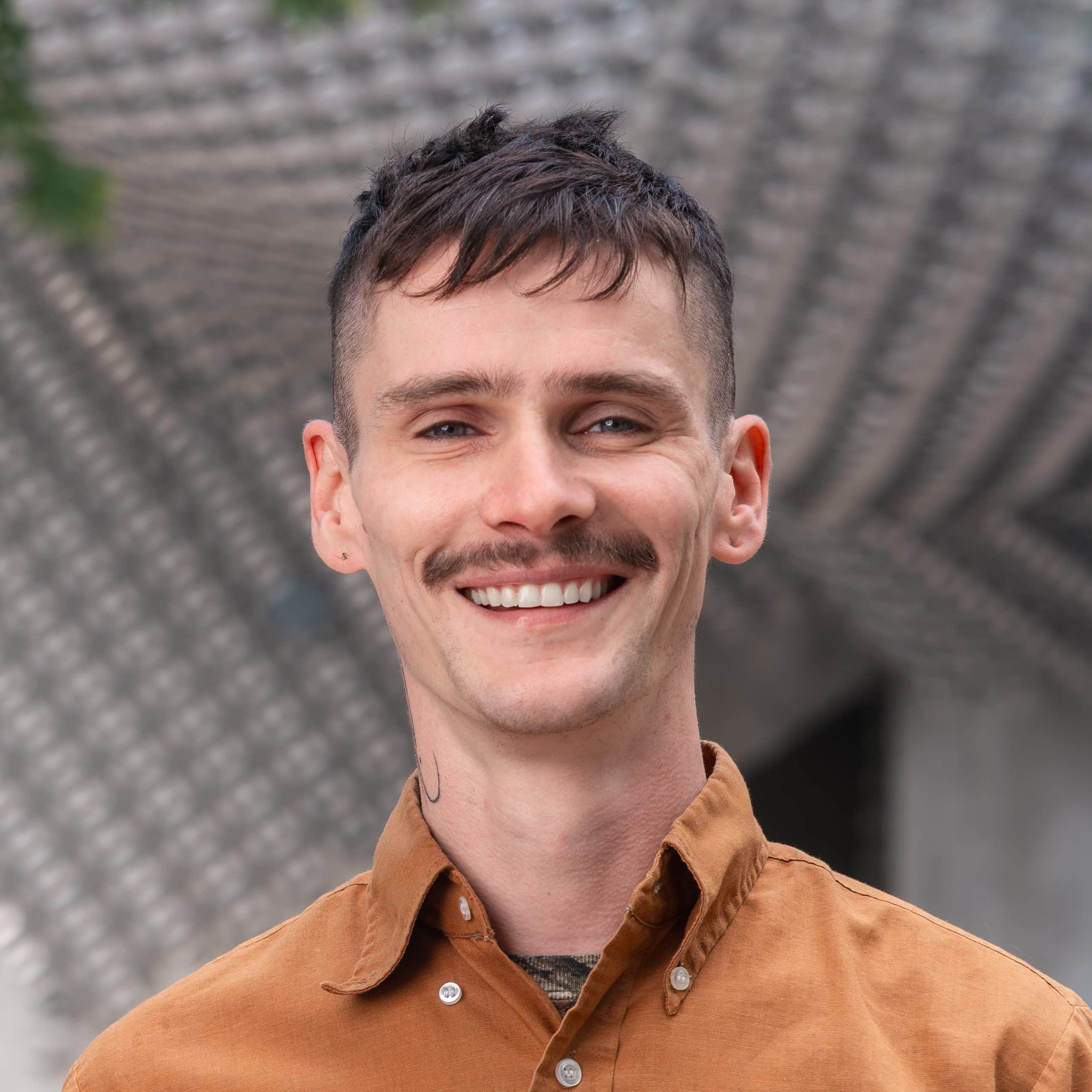 A smiling individual with short hair and a mustache wearing a brown shirt, standing outdoors in an urban setting with green trees and a modern architectural background.