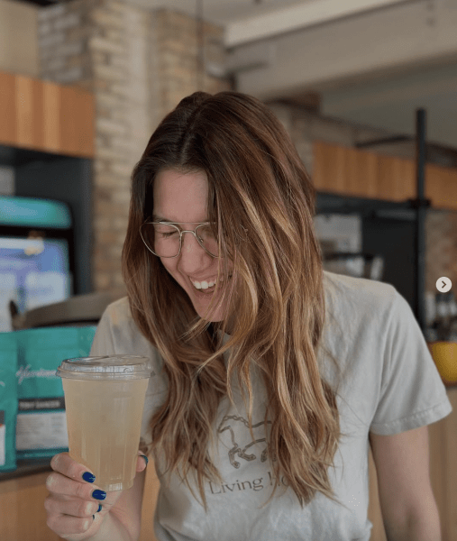 A woman with long hair and glasses is smiling while holding a clear cup with a light-colored drink, standing in a café interior.
