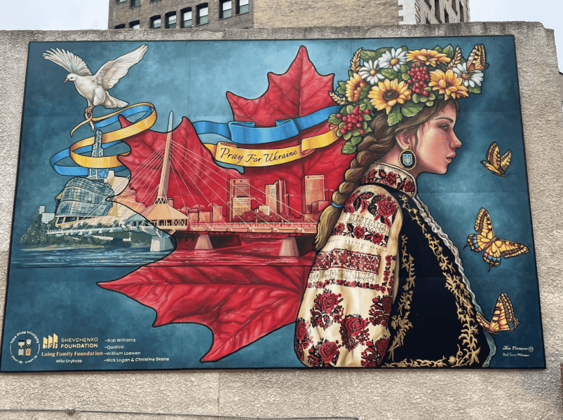 A vibrant mural featuring a young woman with a floral headpiece and traditional Ukrainian attire, set against a backdrop of a maple leaf, city skyline, and a white dove, with the text 'Pray For Ukraine.'