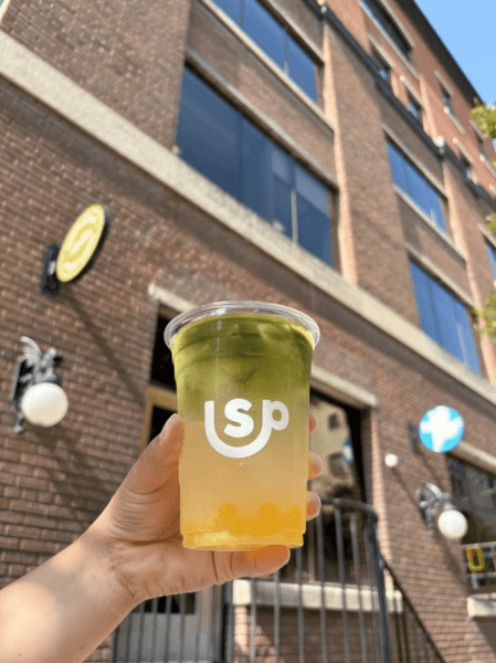 A hand holding a cup of a colorful drink featuring layers of green tea and a yellow base, against the backdrop of a brick building.