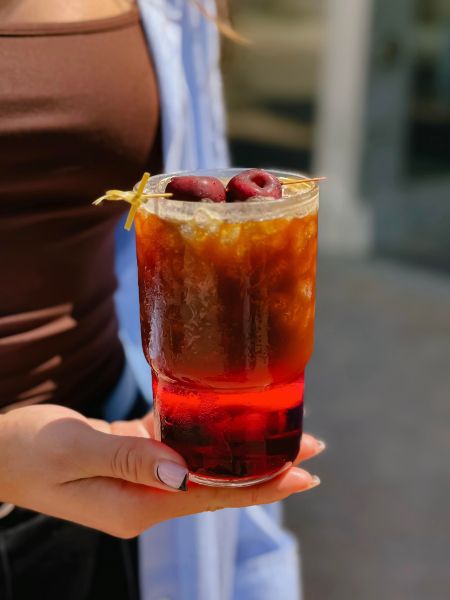 A person holding a refreshing drink in a tall glass, featuring layers of dark and red liquid, topped with cherries and a twist of lemon.