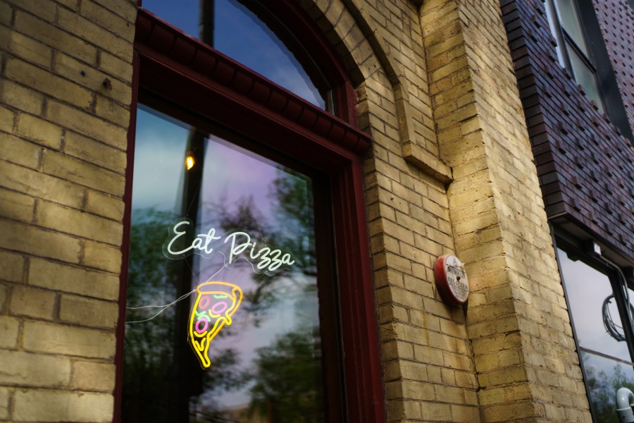 A close-up of a window featuring a neon sign that says 'Eat Pizza' and a colorful pizza slice illustration, set against a brick wall.